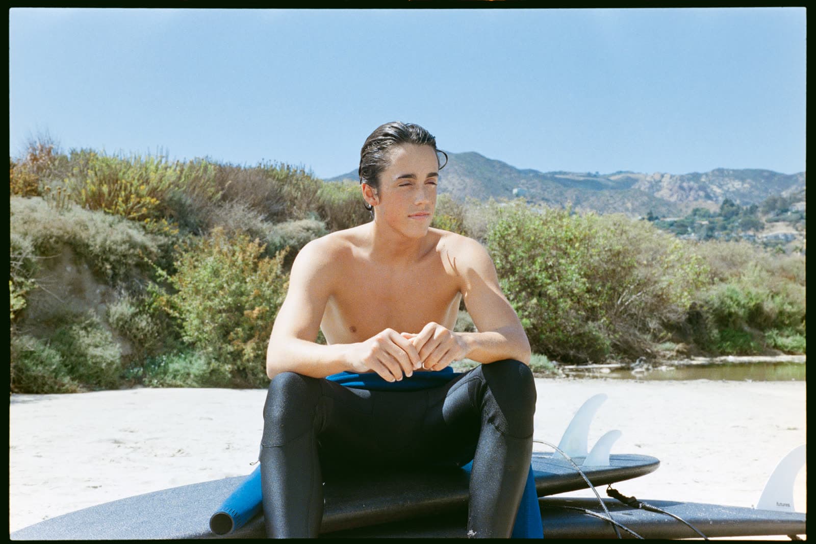 Orion sitting on a surfboard on the beach, hills behind him